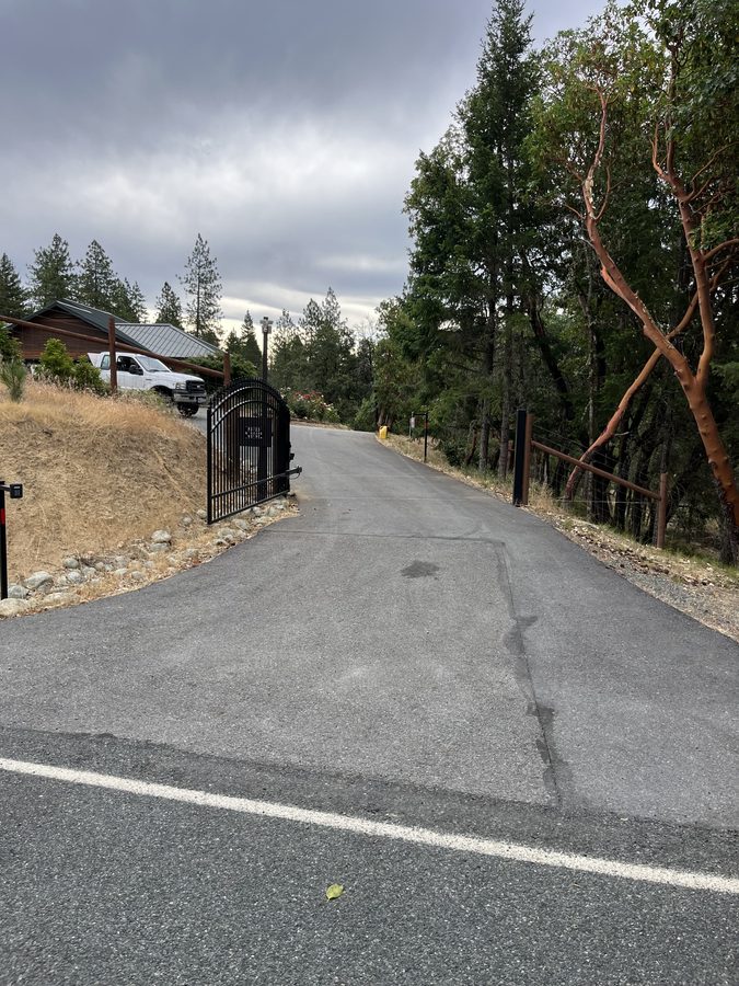 Asphalt driveway with filled cracks and a patched section bordered by a river-rock retaining wall before sealcoating on Seclusion Loop in Grants Pass, Oregon