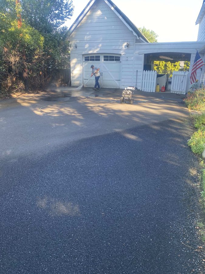 Neill Sealcoating crew member spray-applying fresh black sealcoat across a residential driveway in front of a white home on Hazel Street in Central Point, Oregon